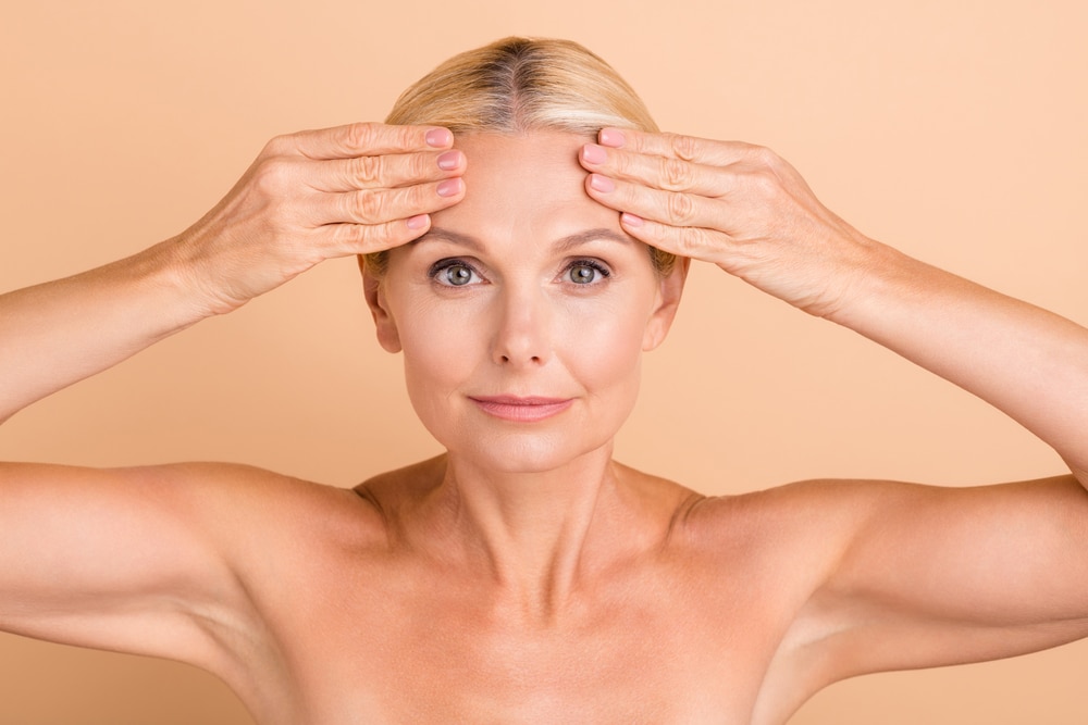 Older woman pulling on her forehead to show her need for a forehead lift to smooth wrinkles.