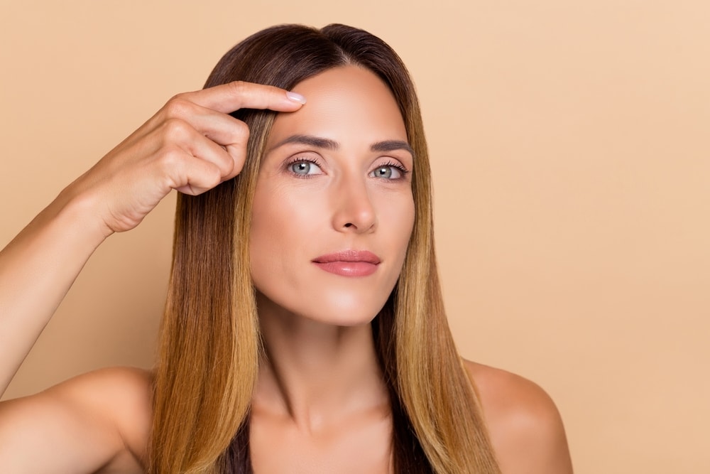 Middle-aged woman touching her brow area in the mirror after receiving a brow lift.